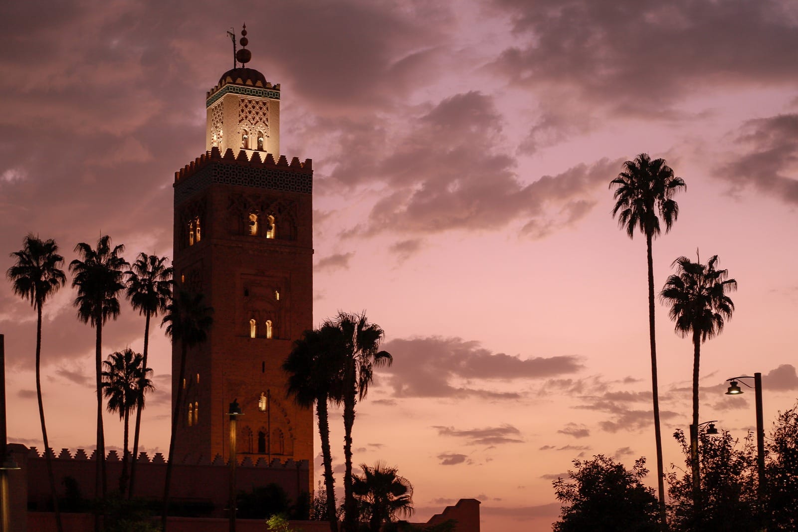 Floodlit Koutoubia minaret behind palm silhouettes under a pink sunset sky